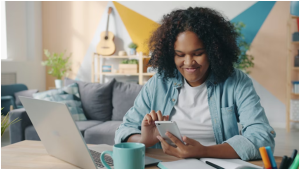 a person using a smartphone at a desk with a laptop. The person appears to be working or studying in a modern office or home setting.