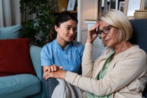 Young woman doctor health care worker medic nurse comforting older senior woman at home visit, patient suffering from clinical depression and anxiety. Sick adult female in home-care program