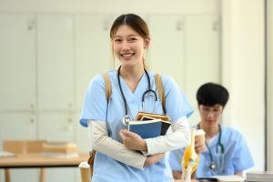 Smiling medical student in blue scrubs carrying textbooks standing in a classroom